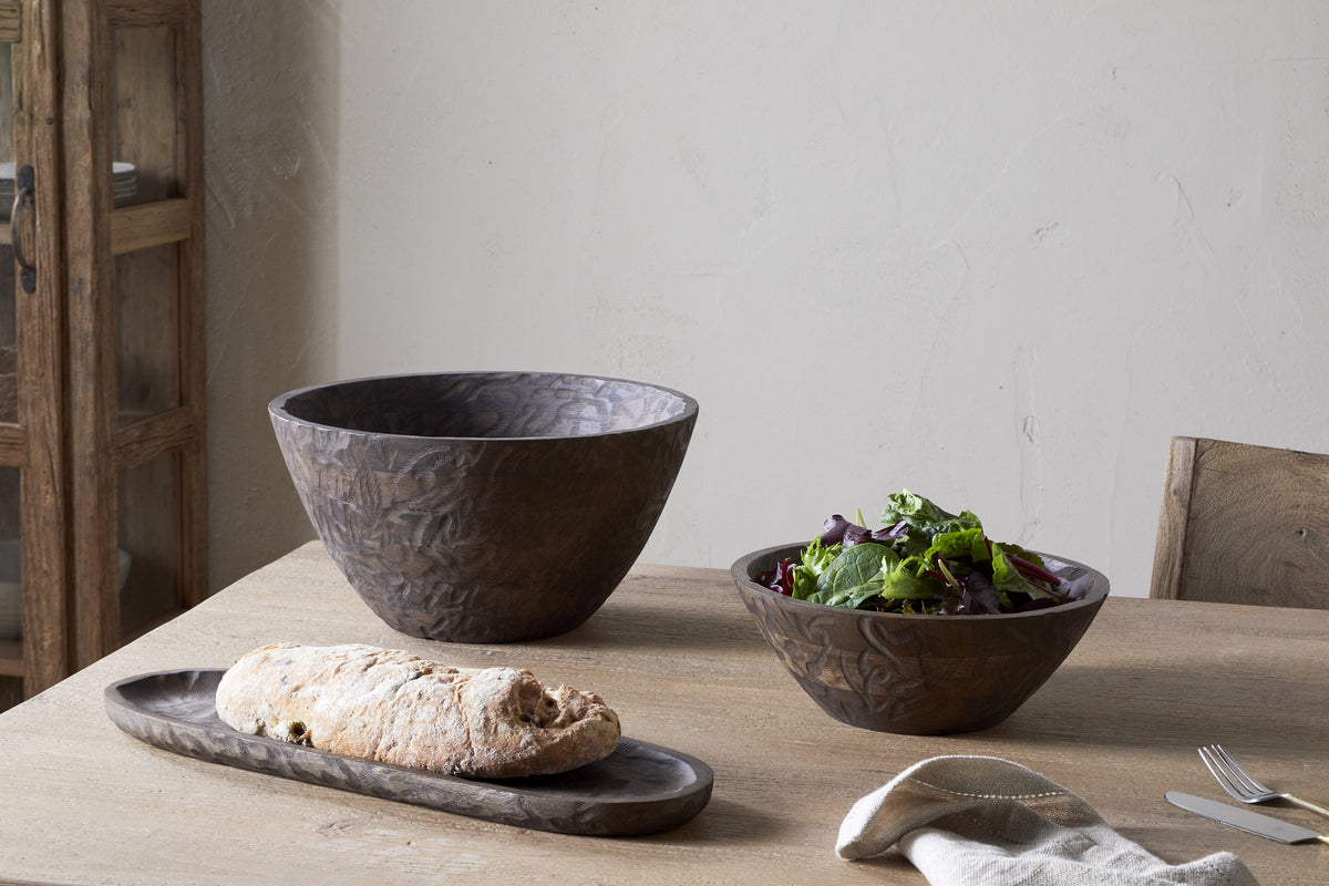 A wooden table set with a long tray holding bread, a medium handcrafted salad bowl, and the nkuku Nyati Wooden Serving Bowl in dark brown (large); utensils and a napkin are nearby.