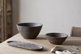 The nkuku Nyati Wooden Serving Bowl - Dark Brown - Large is displayed on a wooden table beside a folded beige napkin, fork, and knife, with a rustic cupboard and textured light wall in the background.