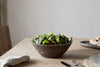 The nkuku Nyati Wooden Serving Bowl - Dark Brown - Small holds a fresh green salad on a wooden dining table, accompanied by a cloth napkin, fork, and a plate with bread.