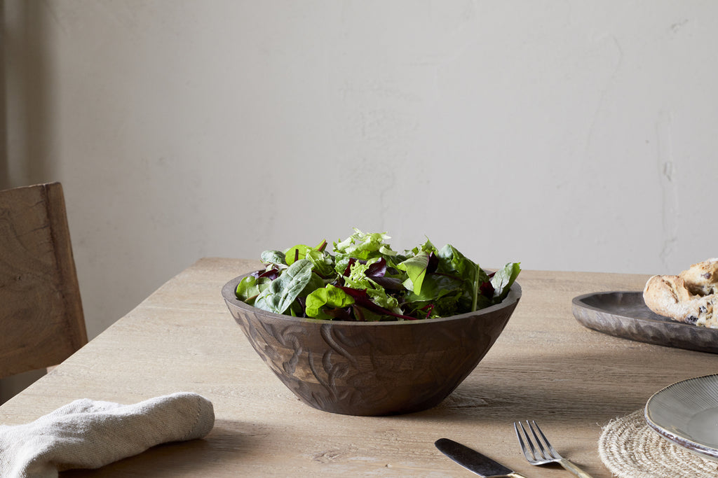 The nkuku Nyati Wooden Serving Bowl - Dark Brown - Small holds a fresh green salad on a wooden dining table, accompanied by a cloth napkin, fork, and a plate with bread.