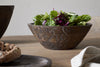 The nkuku Nyati Wooden Serving Bowl - Dark Brown - Small holds a fresh green salad and rests on a wooden table beside a bread loaf and beige napkin. The bowl features carved patterns for added style.