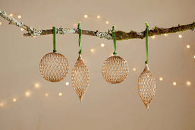 The Nirav Wire Baubles - Brass (Set of 4) from nkuku feature round and elongated brass-finished ornaments, each hung by a green ribbon on a mossy branch, softly glimmering against warm white fairy lights.