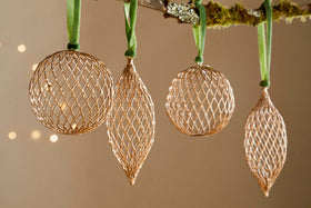 The Nirav Wire Baubles - Brass (Set of 4) by nkuku, featuring three spherical and one elongated bauble, hang from a moss-accented branch with green ribbon. Warm bokeh lights in the background create an elegant festive display.
