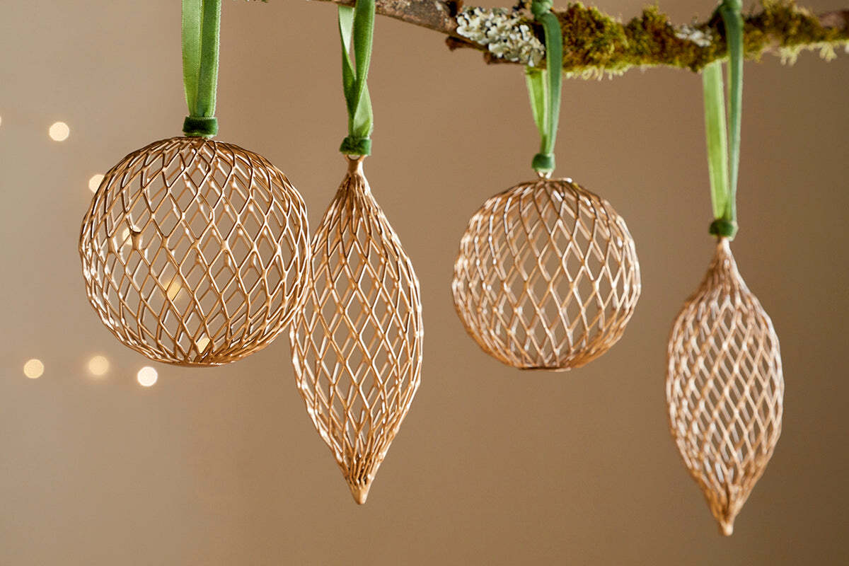 The Nirav Wire Baubles - Brass (Set of 4) by nkuku, featuring three spherical and one elongated bauble, hang from a moss-accented branch with green ribbon. Warm bokeh lights in the background create an elegant festive display.