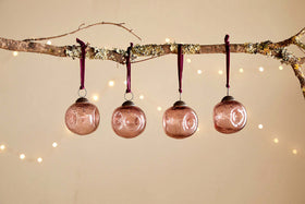 A bare branch displays four nkuku Nishan Pinched Baubles in copper, each hung with a burgundy velvet tie. Warm, blurred lights glow softly against a beige backdrop, adding festive charm to the recycled glass decorations.