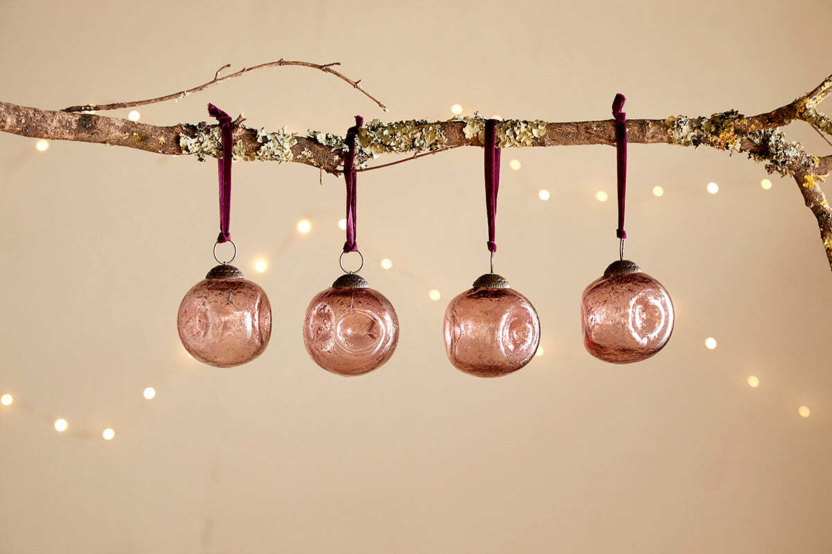 A bare branch displays four nkuku Nishan Pinched Baubles in copper, each hung with a burgundy velvet tie. Warm, blurred lights glow softly against a beige backdrop, adding festive charm to the recycled glass decorations.