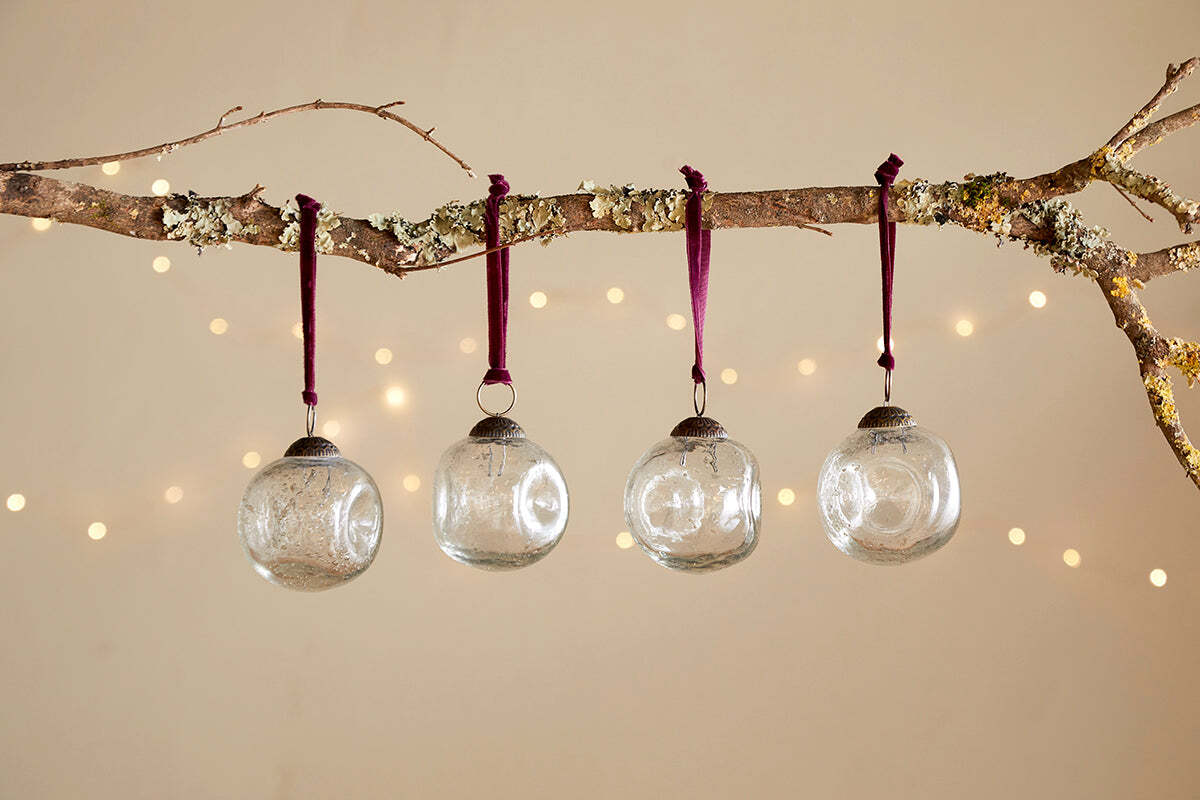 A bare branch with moss and lichen displays four nkuku Nishan Pinched Baubles - Clear, each hung by burgundy velvet ties. Soft fairy lights glow in the background, creating an elegant and festive beige-wall display.