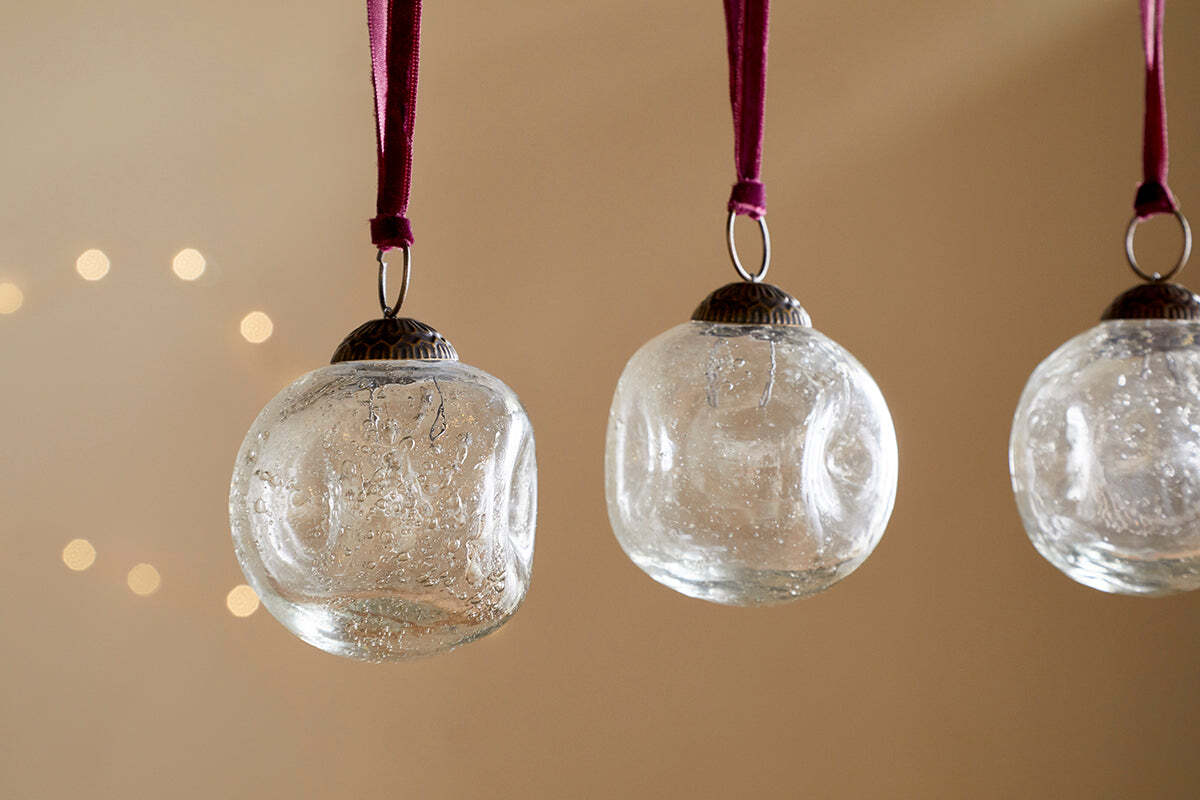 Four clear recycled glass Nishan Pinched Baubles by nkuku hang from burgundy velvet ties against a neutral beige backdrop, with soft, out-of-focus lights creating a warm, festive glow.