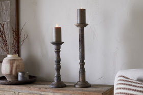 Two lit brown pillar candles stand on nkuku Nayati Rustic Wood Pillar Candle Sticks with a dark stain, each at a different height. They rest on a wooden surface amid rustic decor including a textured vase with dry branches and a candle tray.