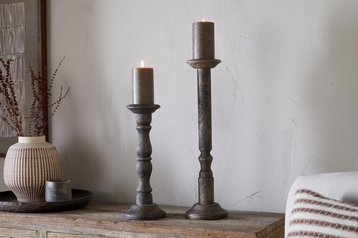 Two lit brown pillar candles stand on nkuku Nayati Rustic Wood Pillar Candle Sticks with a dark stain, each at a different height. They rest on a wooden surface amid rustic decor including a textured vase with dry branches and a candle tray.