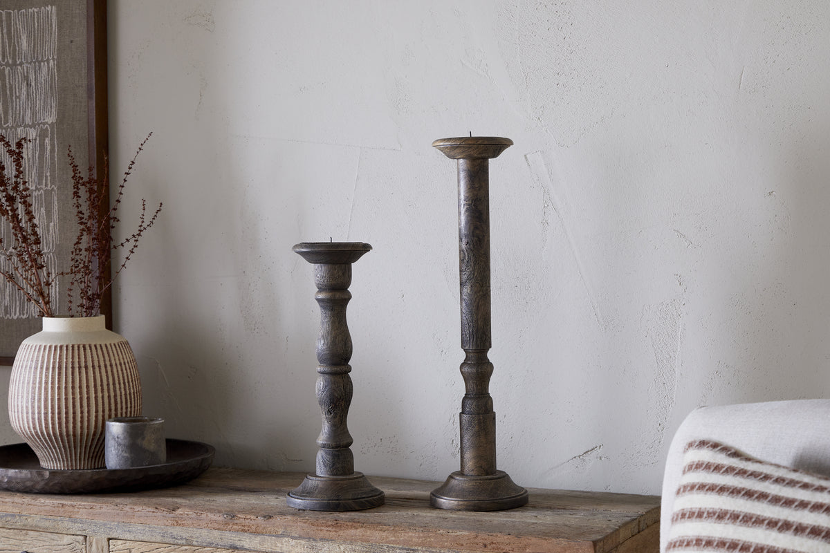 Two Nayati Rustic Wood Pillar Candle Sticks by nkuku in dark stain, each of different heights, stand on a wooden surface beside a ribbed ceramic vase with dried stems and a small metal container on a tray against an off-white textured wall.