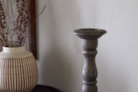 The Nayati Rustic Wood Pillar Candle Stick by nkuku, in a dark stain, stands beside a beige vase with brown stripes and dried branches. A white textured wall behind them adds to the cozy, minimal decor.