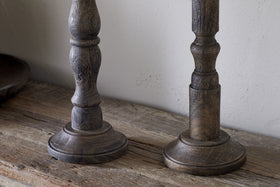 The Nayati Rustic Wood Pillar Candle Stick by nkuku features turned detailing and a dark stain, crafted from sustainable mango wood. Its visible grain and worn texture enhance its vintage charm against a plain, light-colored wall.