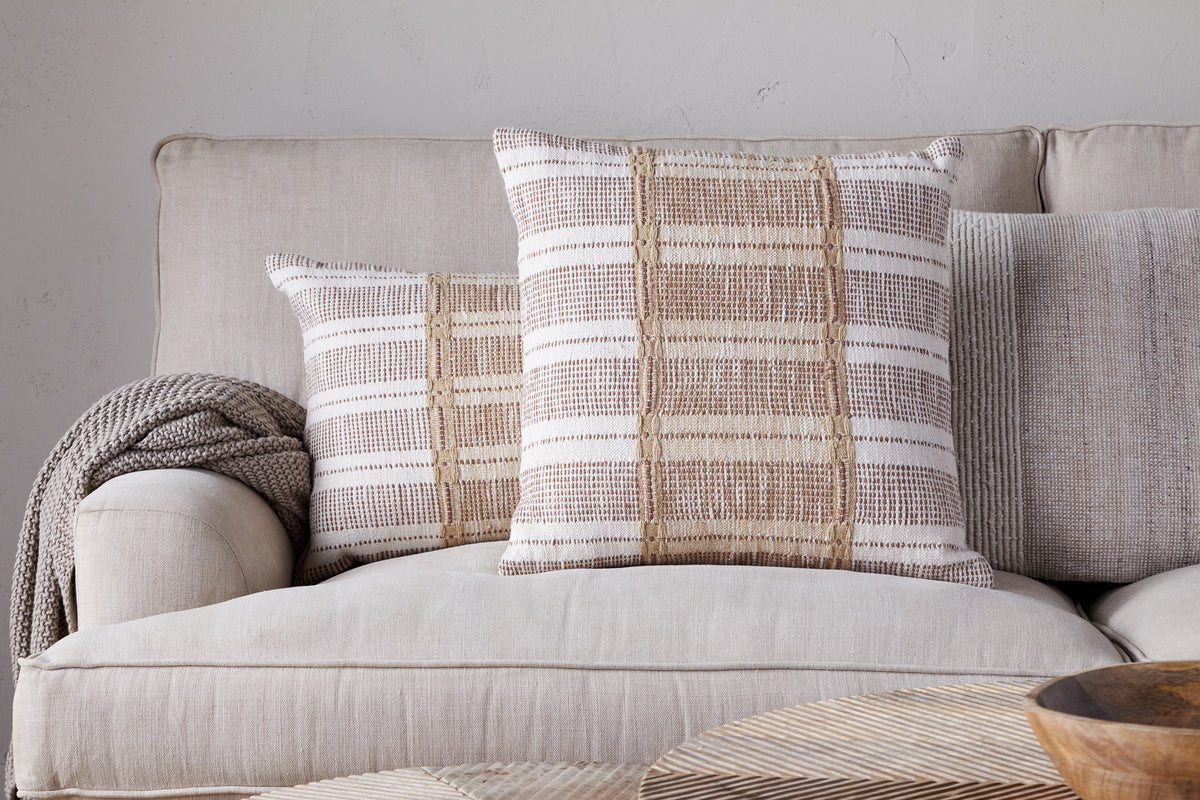 Two Nayika Cotton Cushion Covers by nkuku in rust add warmth to a light tan sofa, with a knitted throw over the armrest. The neutral setting and cosy decor create an inviting atmosphere.