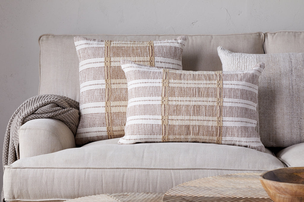 A beige sofa with a cosy cushion and the nkuku Nayika Cotton Cushion Cover in Rust, a knitted gray blanket draped over the armrest, and a wooden tray partially visible in the foreground.