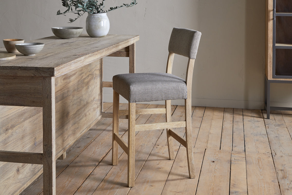 The Nadeeka Linen Upholstered Counter Chair by nkuku, in soft grey, sits beside a rustic wooden table on a light wood floor, topped with ceramic bowls and a vase with greenery—perfect for your kitchen island.