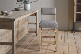 A wooden dining table with ceramic bowls and a white vase sits on a wooden floor next to the nkuku Nadeeka Linen Upholstered Counter Chair in Soft Grey, featuring linen upholstery and a supportive backrest.