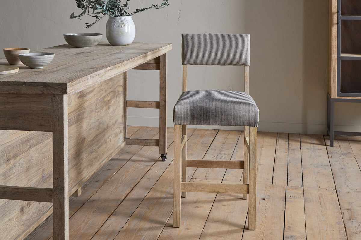A wooden dining table with ceramic bowls and a white vase sits on a wooden floor next to the nkuku Nadeeka Linen Upholstered Counter Chair in Soft Grey, featuring linen upholstery and a supportive backrest.
