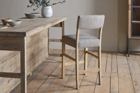 The nkuku Nadeeka Linen Upholstered Counter Chair in Soft Grey, featuring a cushioned backrest, stands by a rustic dining table on a light wood floor holding ceramic bowls and a vase with greenery.
