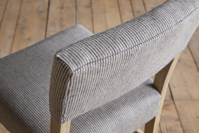 A close-up of the nkuku Nadeeka Linen Upholstered Counter Chair in Soft Grey, featuring striped linen fabric and a wooden frame, shown on a plank wooden floor.