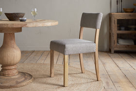 The Nadeeka Linen Upholstered Dining Chair in Soft Grey by nkuku, with a cushioned seat and upholstered backrest, sits on a woven round rug next to a wooden dining table. A wooden sideboard and glasses of white wine are visible in the background.