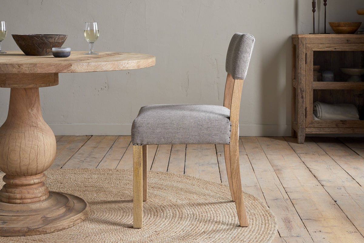 The Nadeeka Linen Upholstered Dining Chair in Soft Grey by nkuku sits on a round woven rug beside a rustic wooden table set with glasses and bowls in a cozy, minimalist dining room.