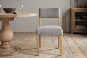 The nkuku Nadeeka Linen Upholstered Dining Chair in Soft Grey, with a cushioned seat, sits on a woven rug beside a round wooden table in a room with light wood floors, neutral walls, and rustic decor.