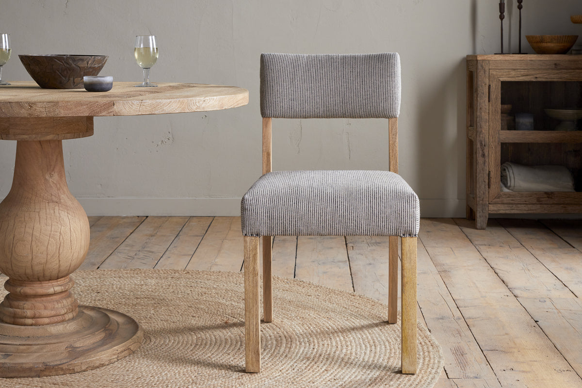 The nkuku Nadeeka Linen Upholstered Dining Chair in Soft Grey, with a cushioned seat, sits on a woven rug beside a round wooden table in a room with light wood floors, neutral walls, and rustic decor.