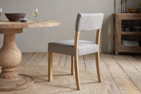 The Nadeeka Linen Upholstered Dining Chair by nkuku in soft grey sits on a round woven rug beside a rustic wooden table set with bowls and wine glasses, in a cozy, light-filled room with wood floors and simple decor.