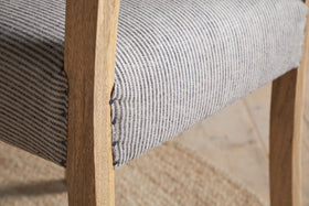 Close-up of the nkuku Nadeeka Linen Upholstered Dining Chair in Soft Grey, featuring a cushioned seat with black stripes, set on a woven beige rug.