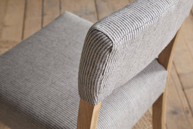 Close-up of the nkuku Nadeeka Linen Upholstered Dining Chair - Soft Grey, with striped linen fabric, cushioned seat and backrest, and light wood legs on a wooden floor.