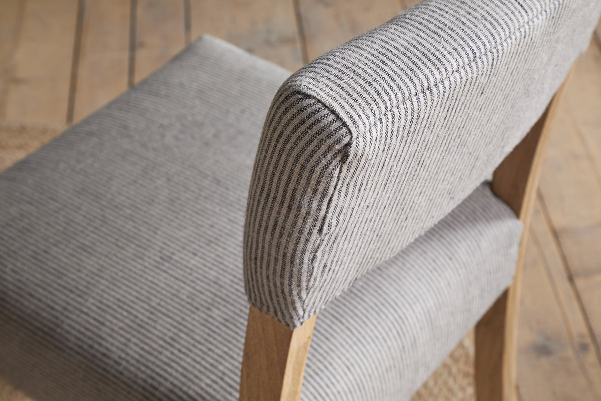 Close-up of the nkuku Nadeeka Linen Upholstered Dining Chair - Soft Grey, with striped linen fabric, cushioned seat and backrest, and light wood legs on a wooden floor.