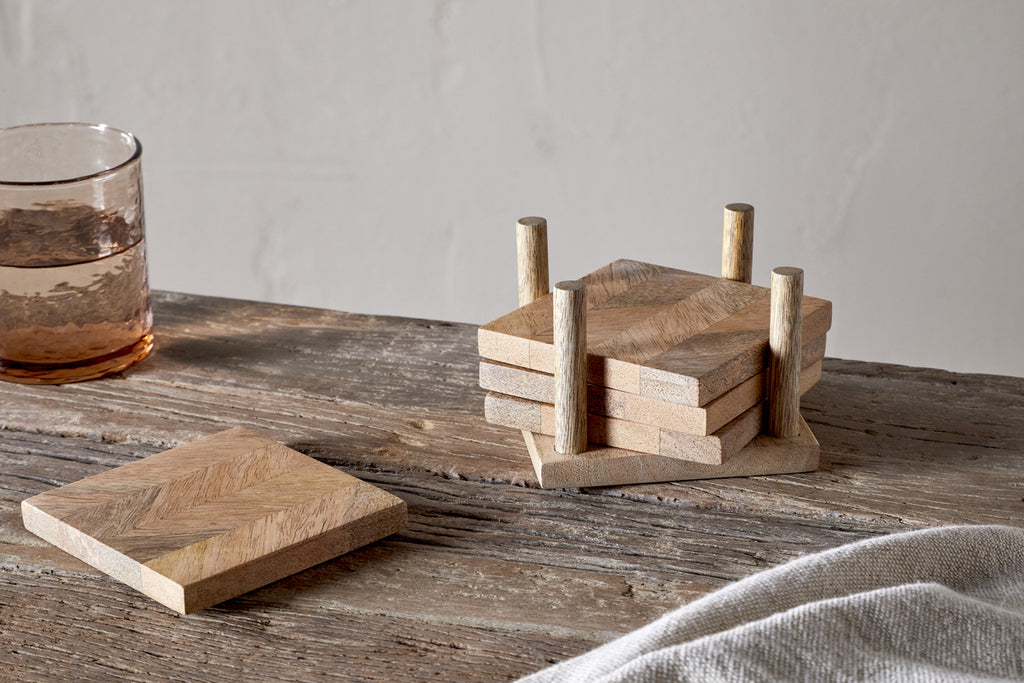 The Nalbari Coasters - Natural (Set of 4) by nkuku are stacked in their holder on a rustic table, with one coaster outside the holder. A glass of water and a light-colored cloth complete the scene.