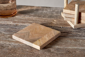 A glass with a brown drink sits on the left, while nkuku’s Nalbari Coasters - Natural (Set of 4), featuring square mango wood and a chevron pattern, rest in their wooden holder on a rustic table.