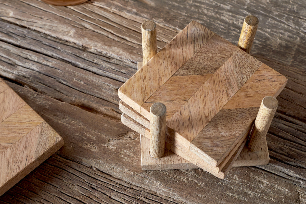 A set of four Nalbari Coasters - Natural by nkuku, featuring a chevron pattern, is stacked in a matching holder on a rustic table, with one coaster placed beside the stack.