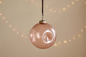 The nkuku Nishan Pinched Giant Bauble - Copper is a large, round, recycled glass ornament with a metallic cap and brown ribbon, featuring a slightly speckled finish. Soft white fairy lights in the background add festive warmth.