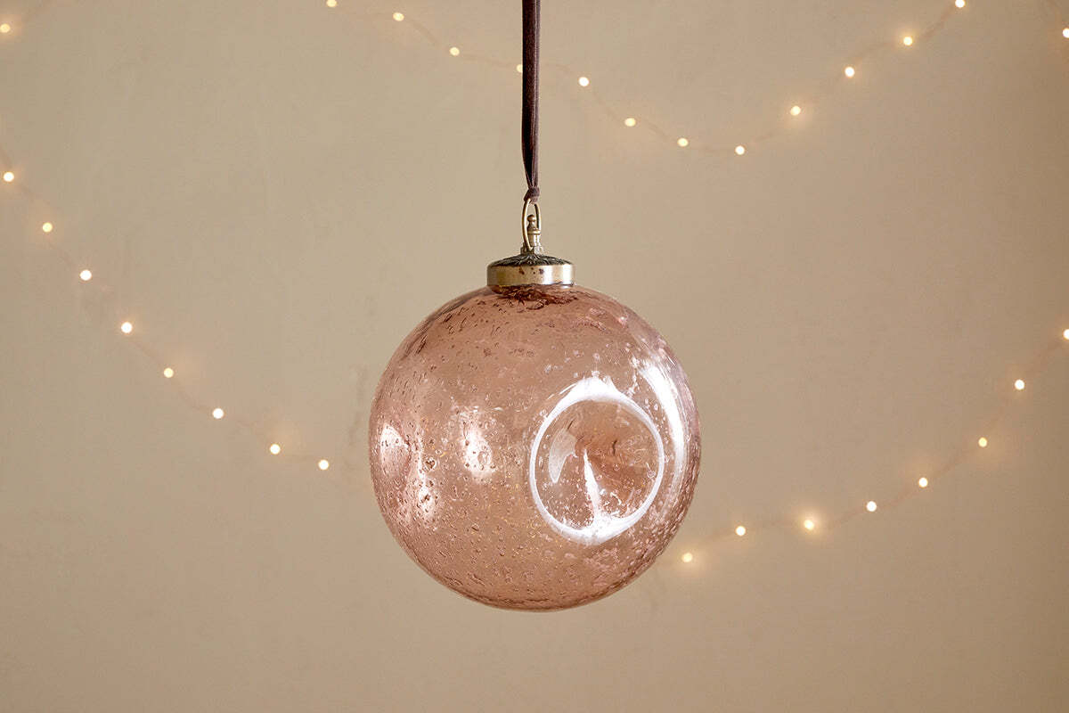 The nkuku Nishan Pinched Giant Bauble - Copper is a large, round, recycled glass ornament with a metallic cap and brown ribbon, featuring a slightly speckled finish. Soft white fairy lights in the background add festive warmth.
