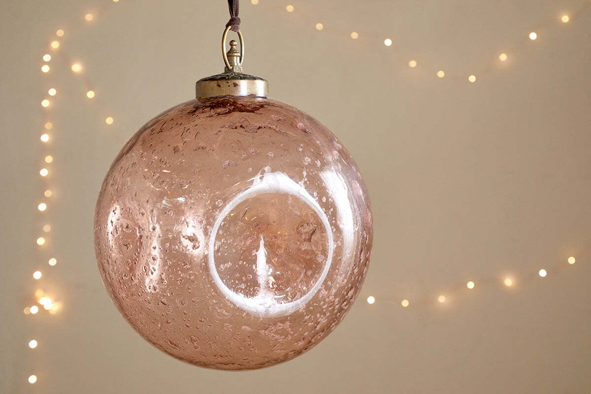 The nkuku Nishan Pinched Giant Bauble - Copper, crafted from recycled glass with beautiful bubbles inside, hangs elegantly from a ribbon. Warm white fairy lights blur in the background, creating a cozy, festive atmosphere as the bauble reflects light.