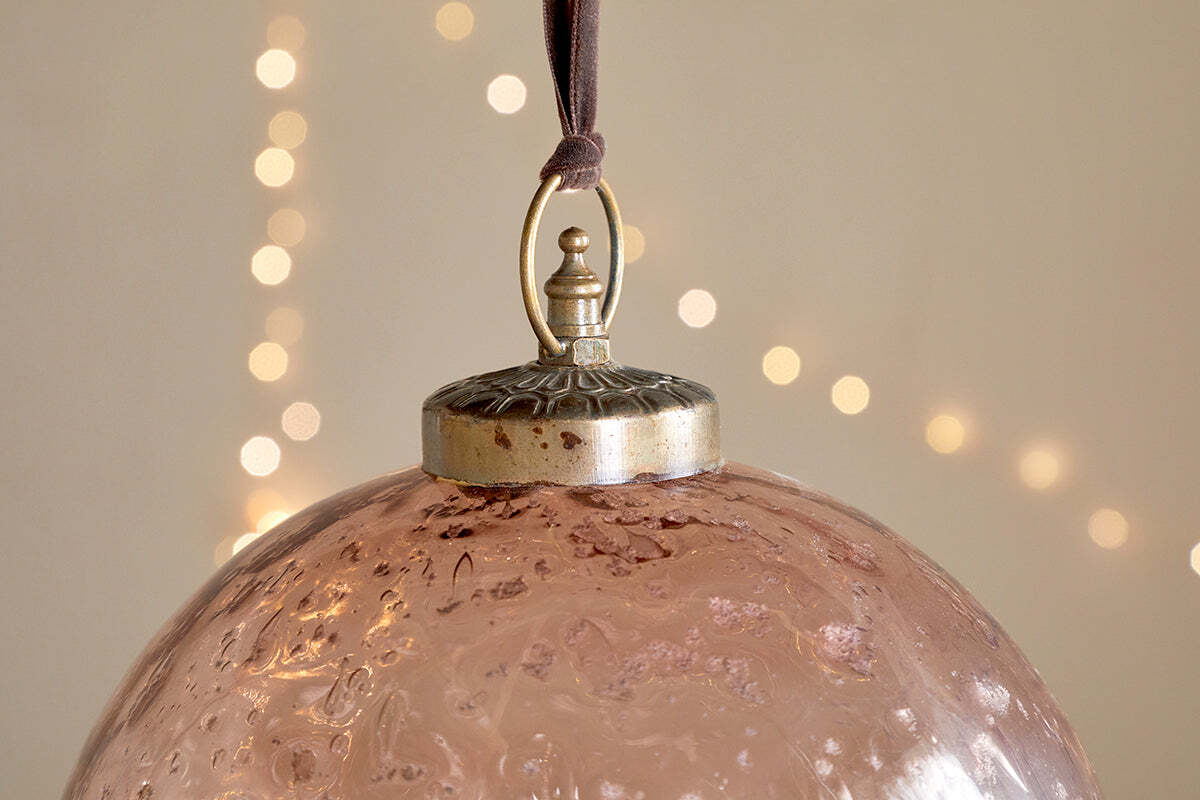 A close-up of nkuku’s Nishan Pinched Giant Bauble - Copper, made from textured recycled glass, hangs by a brown ribbon. Its metallic cap features a decorative loop, with warm white fairy lights softly glowing in the blurred background.