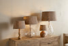 Four nkuku Nadee Banana Leaf Lampshade table lamps, featuring natural textured shades and bases of clear glass and wood, are arranged on a wooden sideboard against a beige wall, creating a cozy, rustic ambiance.