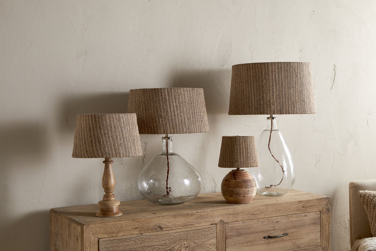 Four Nadee Banana Leaf Lampshades by nkuku, featuring natural textured shades atop glass or wooden bases, are arranged on a rustic wooden sideboard against a neutral wall, creating a cozy and organic look.