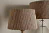 Two nkuku Nadee Banana Leaf Lampshade table lamps, featuring natural textured shades, sit side by side against a plain wall—one with a wooden base in the foreground and one with a glass base in the background.