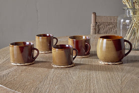 Five Naav Mustard Extra Large Mugs by nkuku rest on woven coasters atop a wooden table. A glass jar with dried plants and a wooden chair with a woven backrest are in the background, completing the scene.
