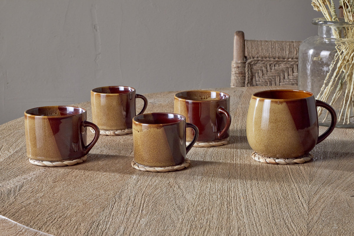 Five Naav Mustard Extra Large Mugs by nkuku rest on woven coasters atop a wooden table. A glass jar with dried plants and a wooden chair with a woven backrest are in the background, completing the scene.