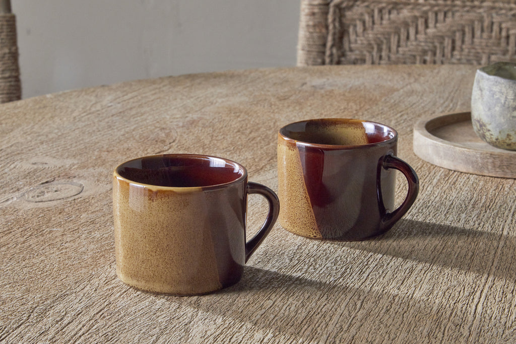 Two Naav Mugs - Mustard - Small (Set of 2) by nkuku sit on a wooden table. Woven chairs and a round tray with a small bowl add to the cozy, softly lit background.