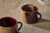 Two Naav Mugs - Mustard - Small (Set of 2) by nkuku, featuring a glossy brown and tan two-tone glaze, rest on a wooden surface. One mug is fully shown; the other appears partially in the lower left, softly lit with gentle shadows.