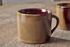 Two Naav Mugs - Mustard - Small by nkuku, featuring a glossy brown finish, are placed on a rustic wooden table; one mug stands in front while part of the second is visible in the background.