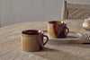 Two Naav Mugs - Mustard (Large, Set of 2) by nkuku rest on a wooden table, one on a tray and the other beside it. A woven chair and small jar in the background enhance the warm, rustic setting.