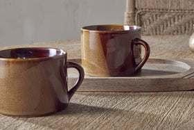 Two Naav Mugs in mustard, large size from nkuku, each with a unique glaze effect, sit on a wooden surface; one is on a round tray and a woven chair is in the background.