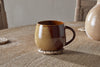 The nkuku Naav Mug - Mustard (Extra Large) features a rounded brown design, resting on a woven coaster on a wooden table, with a ceramic vase and chairs in the background.